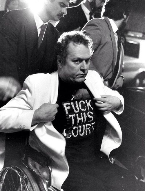 A
    shot of a middle-aged Flynt in a white blazer and black t-shirt that reads,
    Fuck the Court, being wheeled into court.